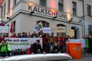 Eine Gruppe von Menschen steht vor einem Gebäude und hält Schilder und Plakate hoch, während zwei Personen im Vordergrund sitzen und ein Müllcontainer rechts zu sehen ist, bei einer Demonstration in Deutschland mit Gebäuden, Fenstern, Lichtern und Schildern im Hintergrund.