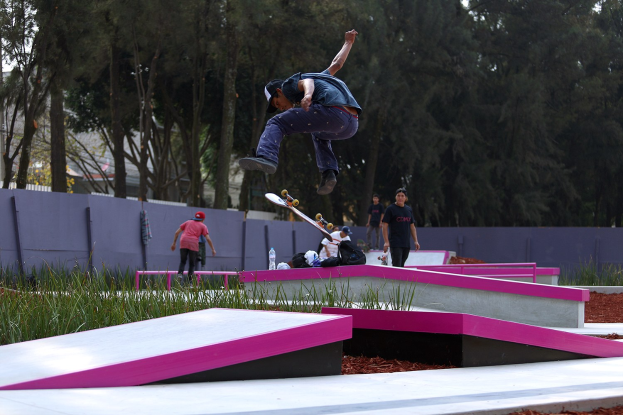 Ein Skateboarder zeigt einen Sprung auf einer Rampe, während Zuschauer zuschauen, mit verstreuten Gegenständen und einem Hintergrund aus Bäumen, Gebäuden und einem klaren Himmel.