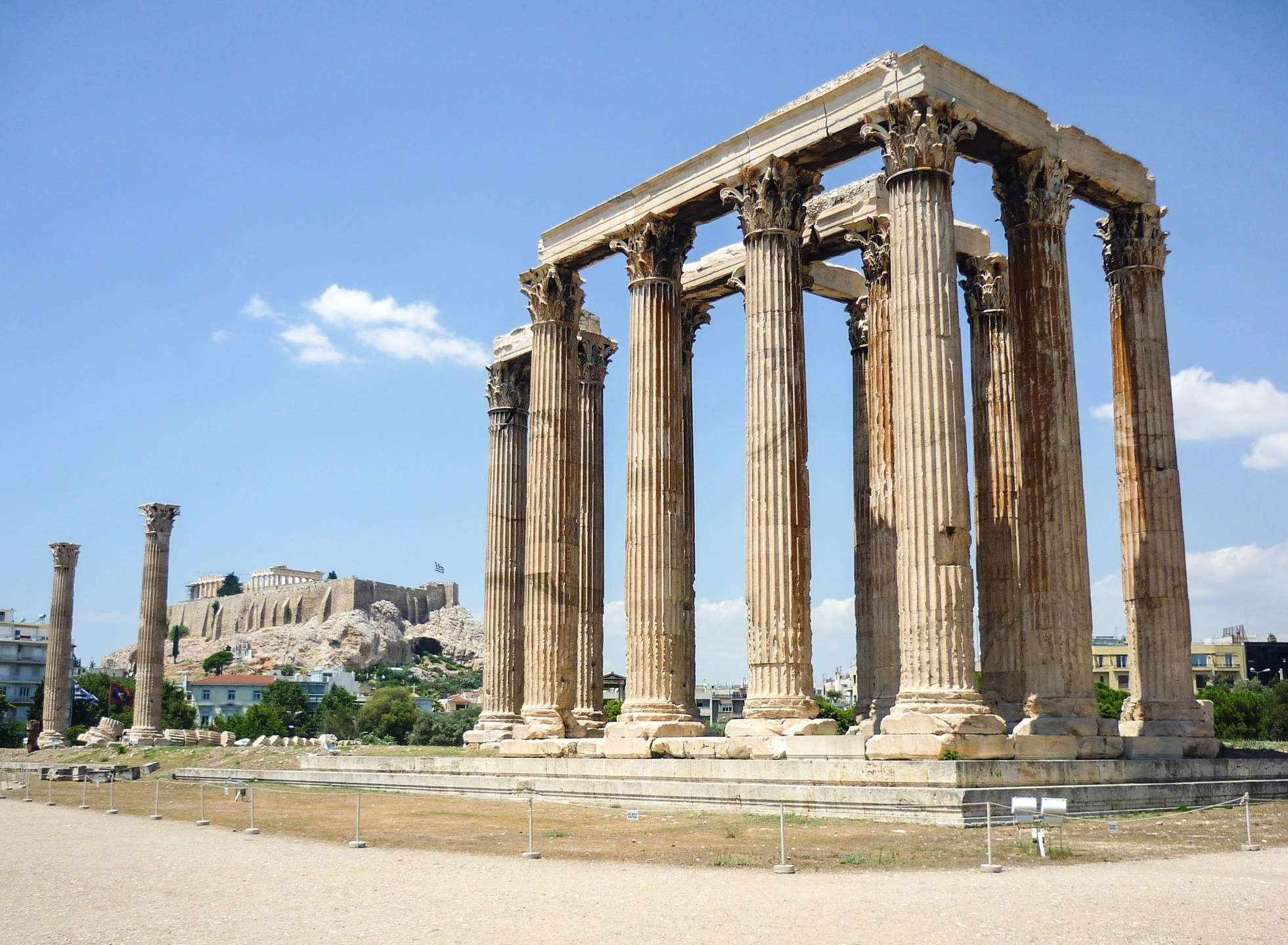Alter Tempel von Olympian Zeus in Athen, Griechenland, mit seinen hohen korinthischen Säulen, umgeben von modernen Gebäuden, Bäumen, steinigem Gelände und einer fernen Festung, unter einem bewölkten Himmel.