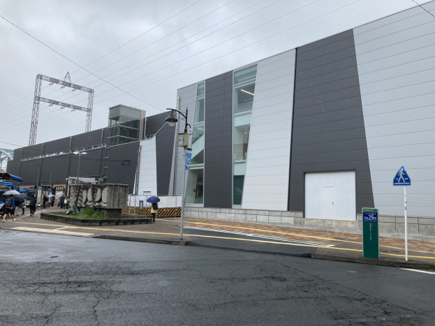 Großes modernes Gebäude mit zahlreichen Fenstern, identifiziert als Tokyo Metropolitan Government Building, umgeben von Straßeninfrastruktur, Fahrzeugen, Fußgängern mit Regenschirmen, Bäumen und einem bewölkten Himmel.