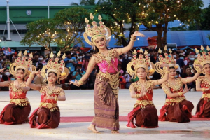 Eine Gruppe von Frauen in traditioneller Thai-Kleidung tanzt vor einem Publikum im Freien, mit Bäumen, Lichtern, Pfählen, einem Gebäude und einem klaren blauen Himmel im Hintergrund.