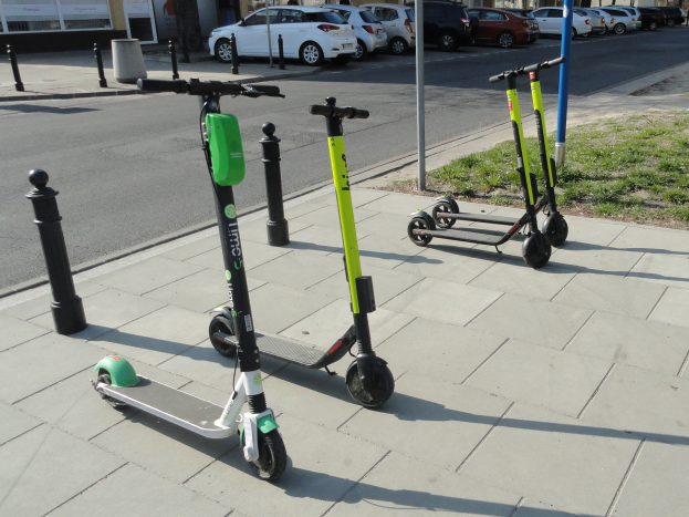 Drei Elektro-Scooter auf einer grasbewachsenen Straßenkreuzung mit Fahrzeugen, Gebäuden, Bäumen und einem klaren Himmel im Hintergrund.