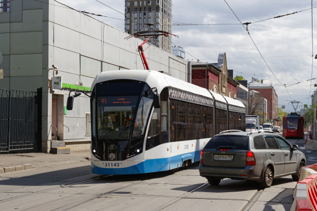 Blauer und weißer Straßenbahnwagen fährt auf einer Stadtstraße mit hohen Gebäuden, Autos, Bäumen, Strommasten und Oberleitungen unter einem bewölkten Himmel.