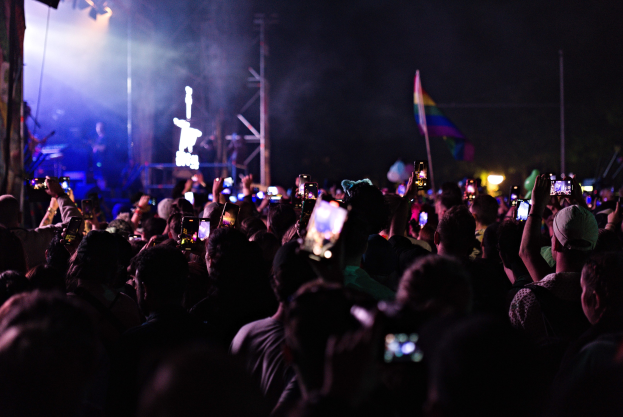 Menge bei einem Konzert mit hochgehaltenen Handys, beleuchtet von Scheinwerfern, umgeben von Pfählen und einer Fahne, während Musiker auf der Bühne spielen.