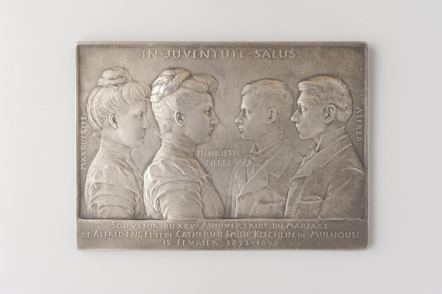 Silberne Plakette mit einem eingravierten Bild von drei Menschen und einem Gedenktext, vor einem weißen Hintergrund.