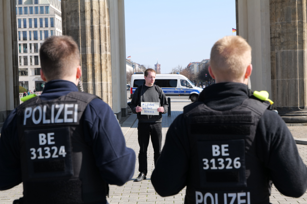 Drei Polizeibeamte stehen vor einem Gebäude mit einem Mann in der Mitte, der ein Papier hält, flankiert von Säulen, mit Fahrzeugen, Menschen, Bäumen, Gebäuden und dem Himmel im Hintergrund.