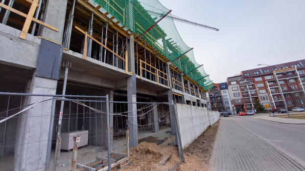 Großer Bau auf einer Stadtstraße mit umliegenden Gebäuden, Straßenlaternen, fahrenden Fahrzeugen und einem klaren blauen Himmel.