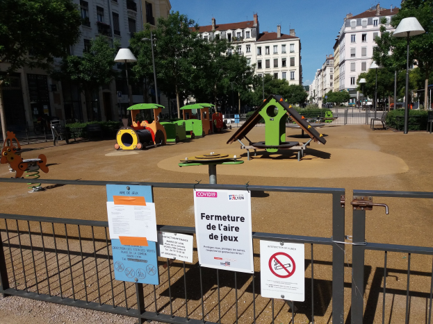 Spielplatz mit einem Zaun und Schildern umgeben, umgeben von Bäumen, Laternenmasten, Gebäuden und einem sichtbaren Himmel auf einem Stadtplatz.