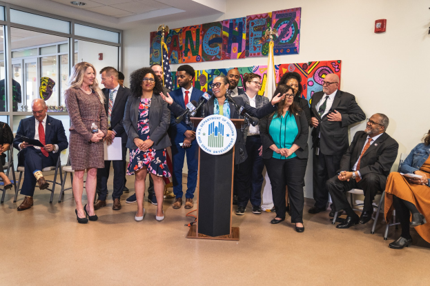 Eine Gruppe von Menschen steht in der Nähe eines Podiums mit einem Mikrofon und einer Tafel mit der Aufschrift "Sancho-Stadtrat tritt dem Stadtrat bei", während andere auf Stühlen sitzen oder in der Nähe stehen; Flaggen, ein Feuerlöscher und Deckenleuchten sind im Hintergrund zu sehen.