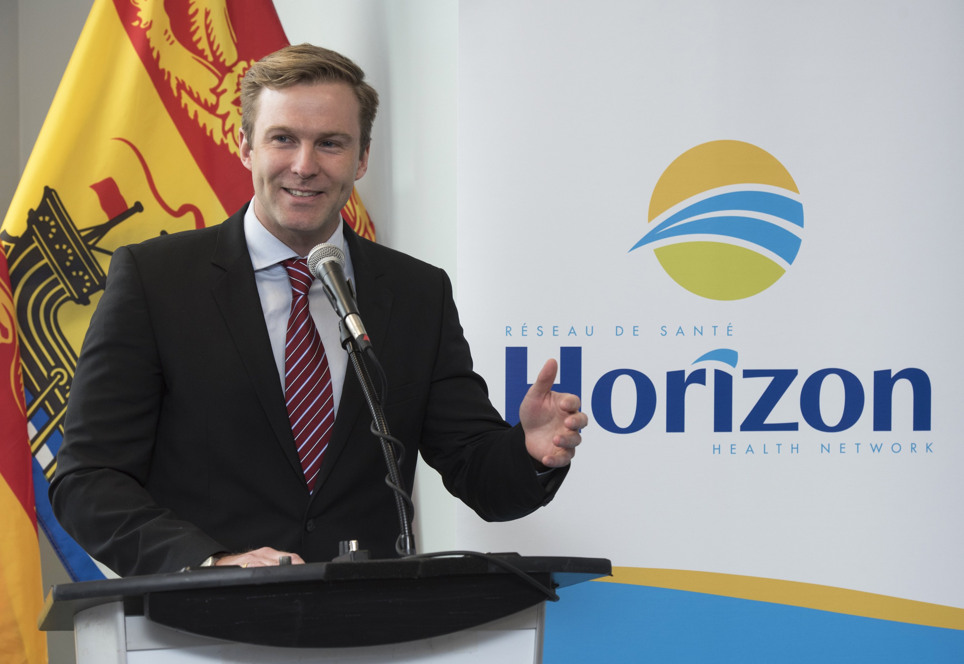 Ein Mann mit einem freundlichen Gesichtsausdruck in einem schwarzen Blazer, einem weißen Hemd und einer roten Krawatte steht an einem Podium mit einem Mikrofon und einem Banner im Hintergrund.