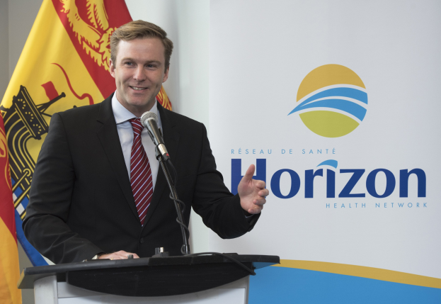 Ein Mann mit einem freundlichen Gesichtsausdruck in einem schwarzen Blazer, einem weißen Hemd und einer roten Krawatte steht an einem Podium mit einem Mikrofon und einem Banner im Hintergrund.