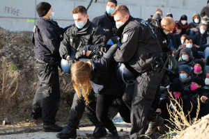 Gruppe maskierter Personen in Kapuzenpullis, einige bewaffnet, die eine Frau umgeben, die von der Polizei eskortiert wird, mit städtischer Vegetation und einem protestbezogenen Wandtext im Hintergrund.