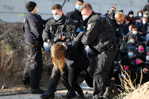 Gruppe maskierter Personen in Kapuzenpullis, einige bewaffnet, die eine Frau umgeben, die von der Polizei eskortiert wird, mit städtischer Vegetation und einem protestbezogenen Wandtext im Hintergrund.