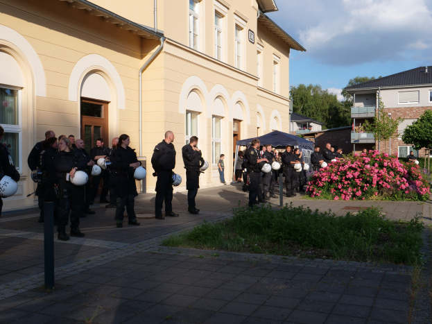 Eine Gruppe von Polizeibeamten vor einem Gebäude mit Fenstern und Türen, die Helme halten, in der Nähe eines Zeltes, umgeben von Pflanzen, Gras, einem Straßenschild, Bäumen und einem bewölkten Himmel.