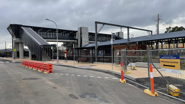 Ein Bahnhof mit einer Brücke im Hintergrund, Straßenlaternen, Strommasten, Stromkabel, Bäume, Gebäude, Fahrzeuge auf der Straße, Absperrgitter, Schilder, Bänke und eine bewölkte Himmel.