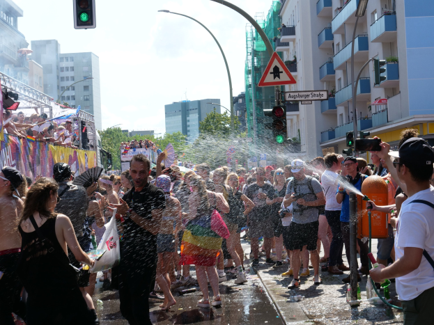 Menschen bei einer Christopher Street Day-Demonstration, die sich gegenseitig mit Wasser bespritzen und Gegenstände halten, mit einem Banner links und Gebäuden, Bäumen und Ampeln im Hintergrund.