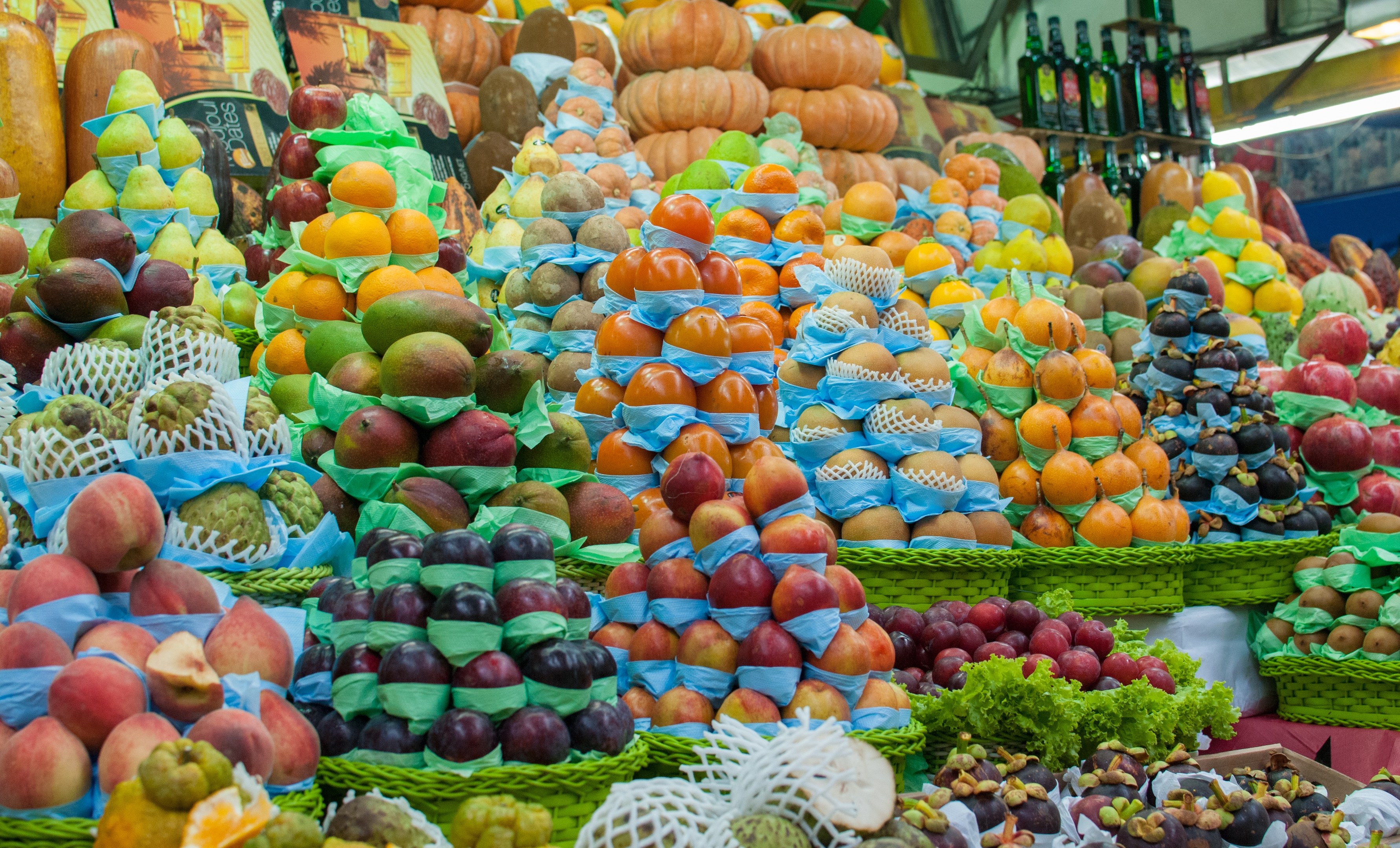 Marktstand mit Äpfeln, Orangen, Birnen und anderen Obst und Gemüse sowie Flaschen auf Regalen und Deckenbeleuchtung.