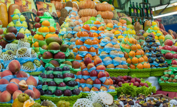 Marktstand mit Äpfeln, Orangen, Birnen und anderen Obst und Gemüse sowie Flaschen auf Regalen und Deckenbeleuchtung.