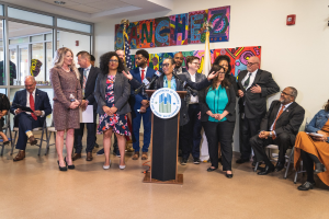 Gruppe von Menschen vor einem Podium mit einem Mikrofon und einer Tafel mit der Aufschrift "Sancho Stadtverordneter tritt dem Stadtrat bei", mit sitzenden und stehenden G├Ąsten, Flaggen, Feuerl├Âscher und Deckenleuchten im Hintergrund.