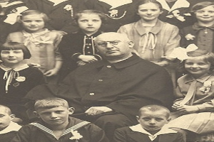 Ein Schwarz-Wei├č-Foto eines ernst dreinschauenden Mannes, der in der Mitte sitzt und von einer Gruppe Kinder umgeben ist, die bewundernd zu ihm aufschauen.