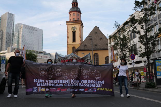 Eine Gruppe von Menschen in Masken, die eine Straße entlanggehen und ein Banner halten, mit einem geparkten Auto auf der linken Seite, Gebäuden, Bäumen, Schildern, Pfosten, einem Uhrenturm und einem klaren blauen Himmel im Hintergrund.