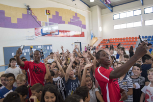 Kinder vor einem Basketballfeld an einem Camp stehend, einige halten Telefone, mit einer Pinnwand, Uhr, Torpfosten, Basketballkorb, Deckenbeleuchtung, Stühlen und Fenstern im Hintergrund.