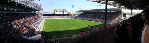 Großes Stadion voller Zuschauer bei einem Fußballspiel, Spieler auf dem Feld, Fahnen, Gebäude, Laternenpfähle, Bäume und einen klaren blauen Himmel im Hintergrund.