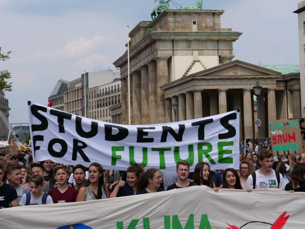 Gruppe von Studenten marschiert in Berlin mit einer bunten "Students for Future"-Fahne vor einer Kulisse aus Gebäuden, Bäumen und Himmel.
