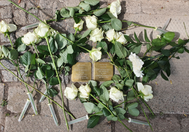 Ein Kranz aus weißen Rosen und Kerzen vor einem Denkmal in Berlin, Deutschland, mit zwei goldfarbenen, beschrifteten Gegenständen.