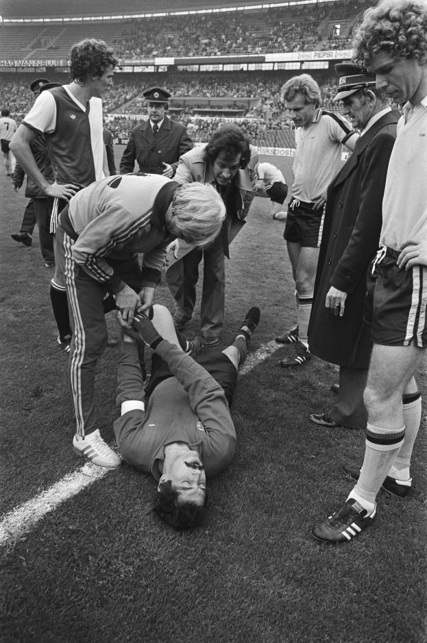 Schwarzes und weißes Bild eines Fußballspielers auf dem Boden umgeben von einer Gruppe von Männern, mit einer Menge im Hintergrund eines Stadions.