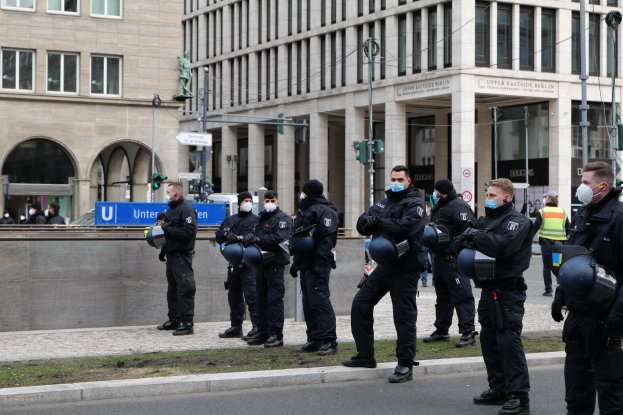 Gruppe von Polizisten in schwarzen Uniformen und Masken vor einem Berliner Gebäude mit Glasfenstern und Säulen, einige halten Helme, mit Laternenmasten, Verkehrsampeln, Informationsschildern, einer Statue und anderen Menschen im Hintergrund auf einem grasbewachsenen Boden.