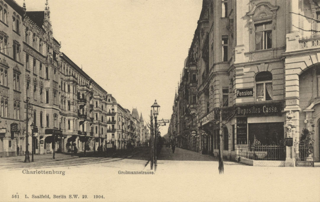 Schwarzes und weißes Foto einer Stadtstraße in Charlottenburg, Deutschland, mit Gebäuden, Straßenmasten, Laternen, Namensschildern, Kraftfahrzeugen, Fußgängern, Gittern und einem klaren Himmel.