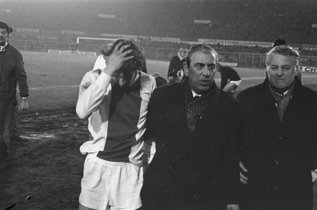 Schwarzes und weißes Bild einer Gruppe von Männern auf einem Fußballfeld mit einem Stadion im Hintergrund, die nach einem Sieg betrübt aussehen.