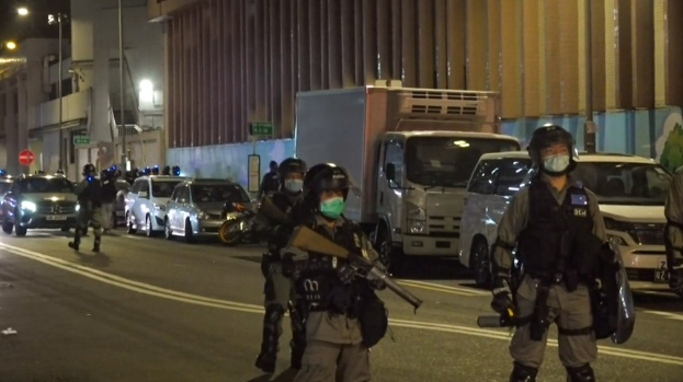 Eine Gruppe von Polizisten in Helmen, Handschuhen und Gesichtsmasken steht an einer Straße in der Nacht, einige halten Waffen, mit Fahrzeugen, Laternen, Schildern und Gebäuden im Hintergrund.