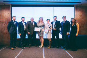 Gruppe von lächelnden Menschen mit Urkunden vor einem Bildschirm, mit einem Tisch und einem Wandobjekt im Hintergrund, Gewinner der Internationalen Geschäftsauszeichnungen 2018.