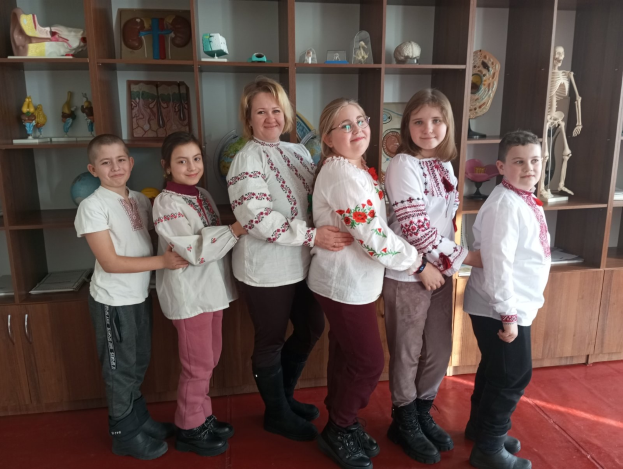 Eine Gruppe von Kindern in traditioneller ukrainischer Kleidung vor einem Bücherregal mit Spielzeug, einem Skelett und anderen Gegenständen, das wie eine ukrainische Schule aussieht.