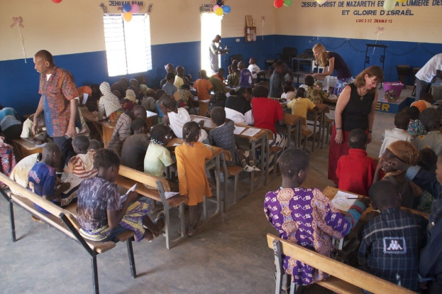 Kinder auf Bänken im Vordergrund mit Erwachsenen, die sie führen, Stühle, eine Tür, ein Fenster, ein Tisch, Ballons und im Hintergrund sichtbarer Text.
