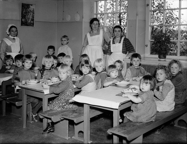 Kinder sitzen an Tischen mit Tellern voller Essen in einem Klassenzimmer, mit Bänken um die Tische und einem Foto Rahmen an der Wand im Hintergrund, Fenster mit Pflanzen draußen rechts.