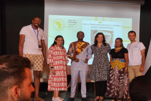 Eine Gruppe von Menschen auf einer Bühne, die lächeln und für ein Foto posieren, mit einem Publikum auf Stühlen und einem Bildschirm im Hintergrund, der 'Open the Knowledge Journalism Awards 2019' anzeigt.