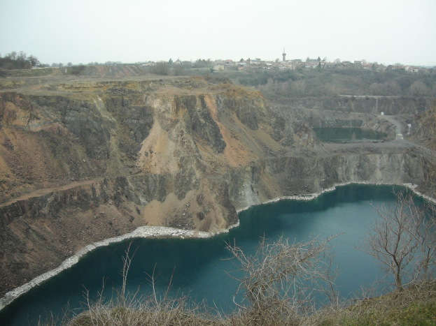 Großer Tagebau mit einem zentralen See, umgeben von Bäumen und Gebäuden unter einem klaren blauen Himmel.