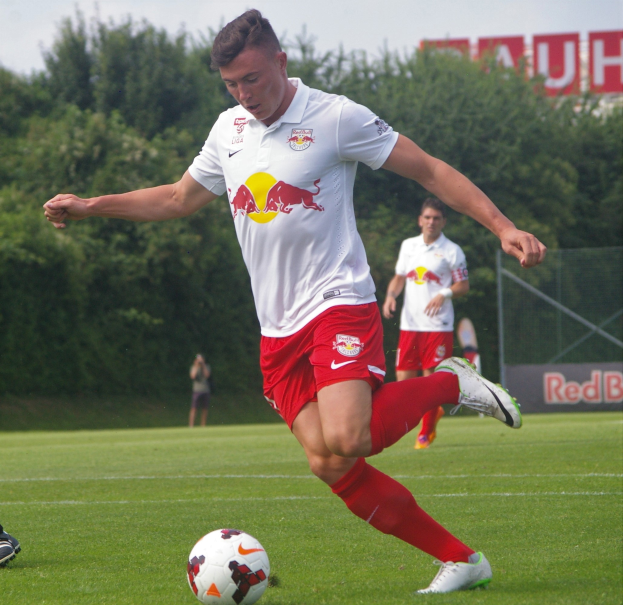 Ein Mann in Fußballkleidung kickt einen Ball auf einem grünen Feld mit Bäumen und einem klaren blauen Himmel im Hintergrund, wobei ein Schild mit der Aufschrift "RB Leipzig" zu sehen ist.