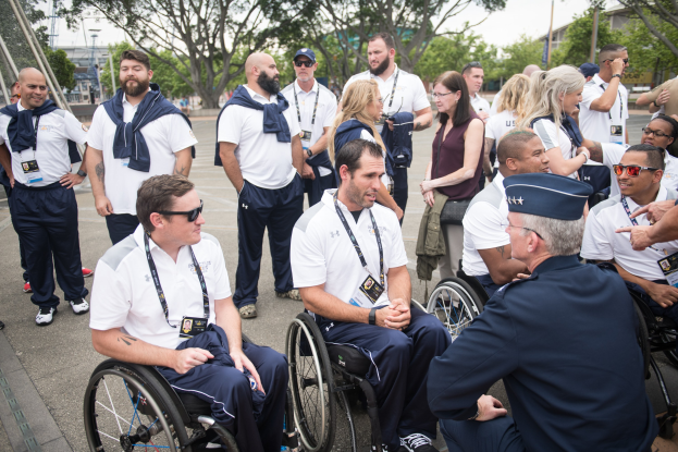 Eine Gruppe von Menschen im Rollstuhl vor einem Hintergrund aus Bäumen, Pfählen, Gebäuden und einem klaren blauen Himmel, wobei einige Mützen und Schutzbrillen tragen, was darauf hindeutet, dass sie bei den Paralympischen Spielen sind.