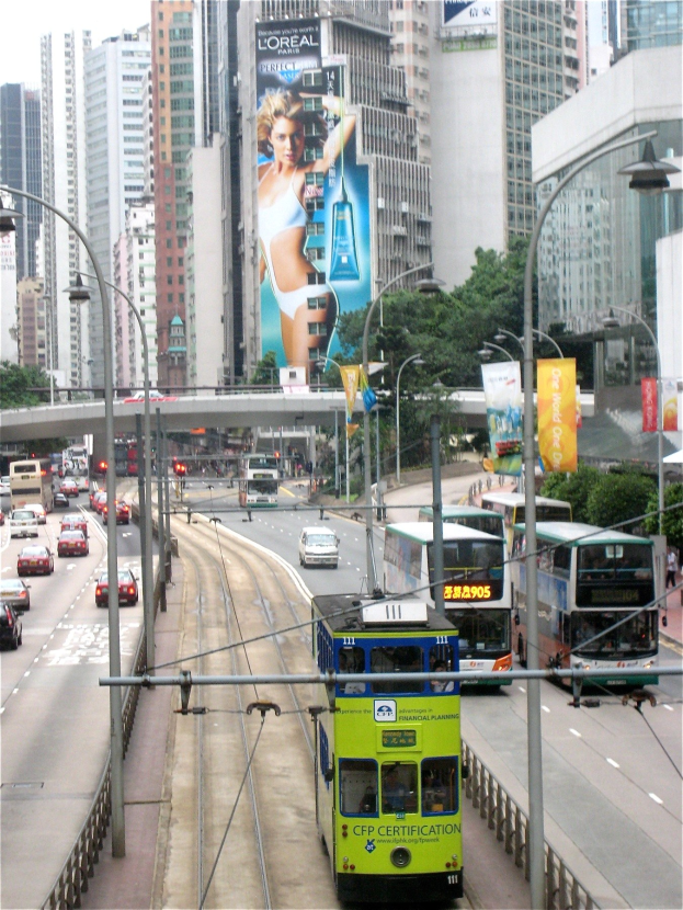 Eine belebte Stadtstraße mit verschiedenen Fahrzeugen, darunter Doppeldeckerbusse, Autos und andere Fahrzeuge, unter einem klaren blauen Himmel, mit hohen Gebäuden, einer Brücke und Schildern im Hintergrund.
