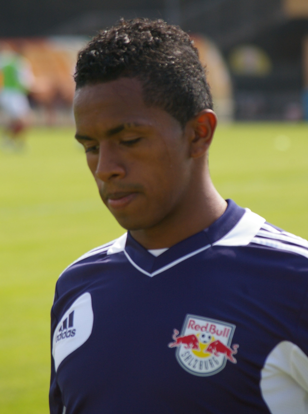 Ein junger Mann in einem blauen T-Shirt steht auf einem Fußballfeld mit einem unscharfen Hintergrund, der als Flügelspieler mit kurzen dunklen Haaren und einem entschlossenen Gesichtsausdruck erscheint.