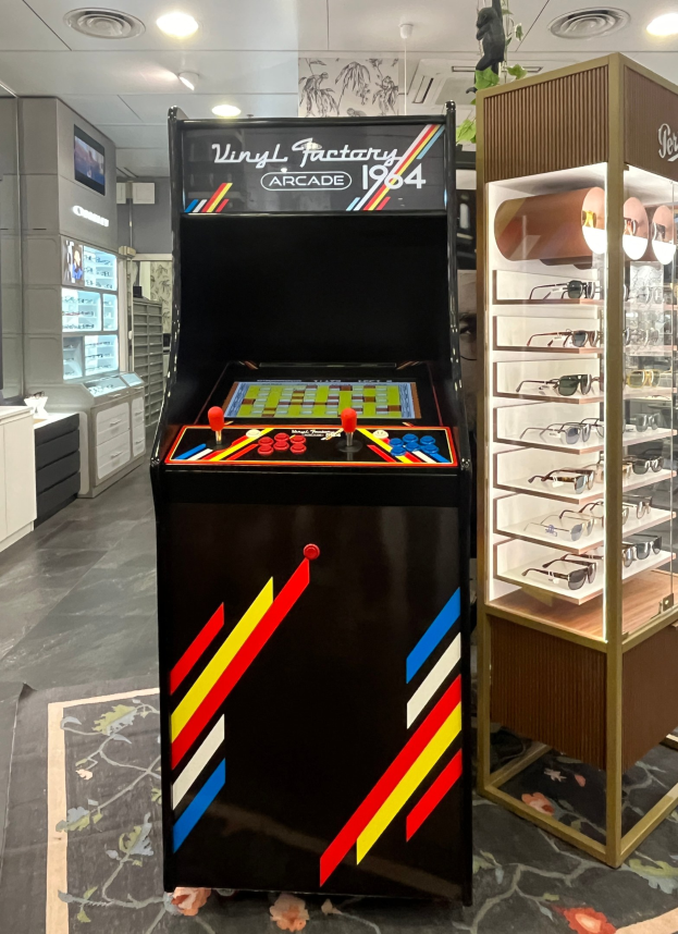 Ein Geschäft mit einem zentralen Retro-Arcade-Spielautomaten, einer Glasvitrine auf der rechten Seite und einer retro-inspirierten Beleuchtung mit Schränken und einem Fernseher im Hintergrund.