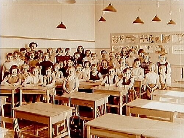 Ein Schwarz-Weiß-Foto einer Klasse mit Kindern auf Bänken und Stehplätzen, umgeben von verstreuten Tischen und Stühlen, mit einer Tafel an der Wand und Deckenleuchten.