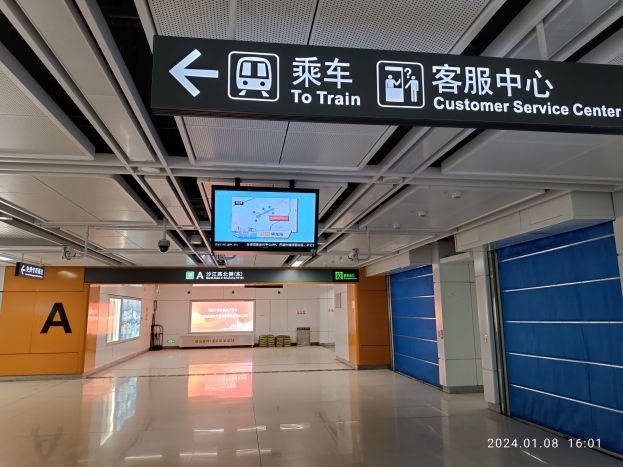 Innenansicht eines Bahnhofs mit einem "Zum Bahnkunden-Servicecenter"-Schild oben, sichtbarem Boden, Türen auf der rechten Seite, deckenmontierten Tafeln, einem zentralen Bildschirm und einem Wasserzeichen unten rechts.