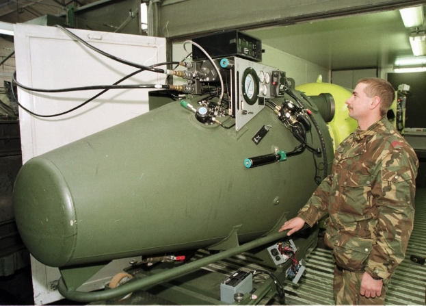 Ein Mann in militärischer Uniform steht neben einer großen grünen Bombe in einem unordentlichen Raum mit Maschinen, Drähten und einer beleuchteten Wand im Hintergrund.