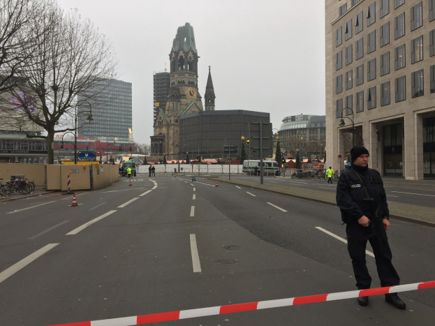 Ein Polizist in schwarzer Uniform und Mütze steht auf einer Straße in Berlin umgeben von Verkehrskegeln, Fahrrädern, Laternenmasten, Bäumen und Gebäuden, im Hintergrund ein Kirchturm und ein blauer Himmel.