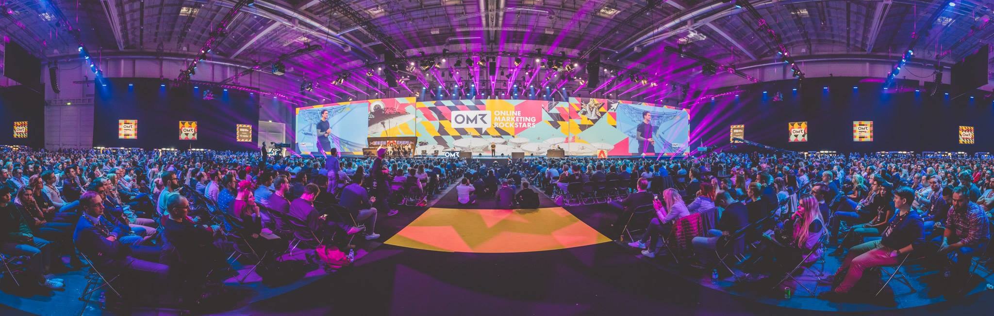 Eine große Zuschauermenge sitzt einer hell erleuchteten Bühne mit Bildschirmen gegenüber, auf der mehrere Menschen während eines Online-Marketing-Events stehen.